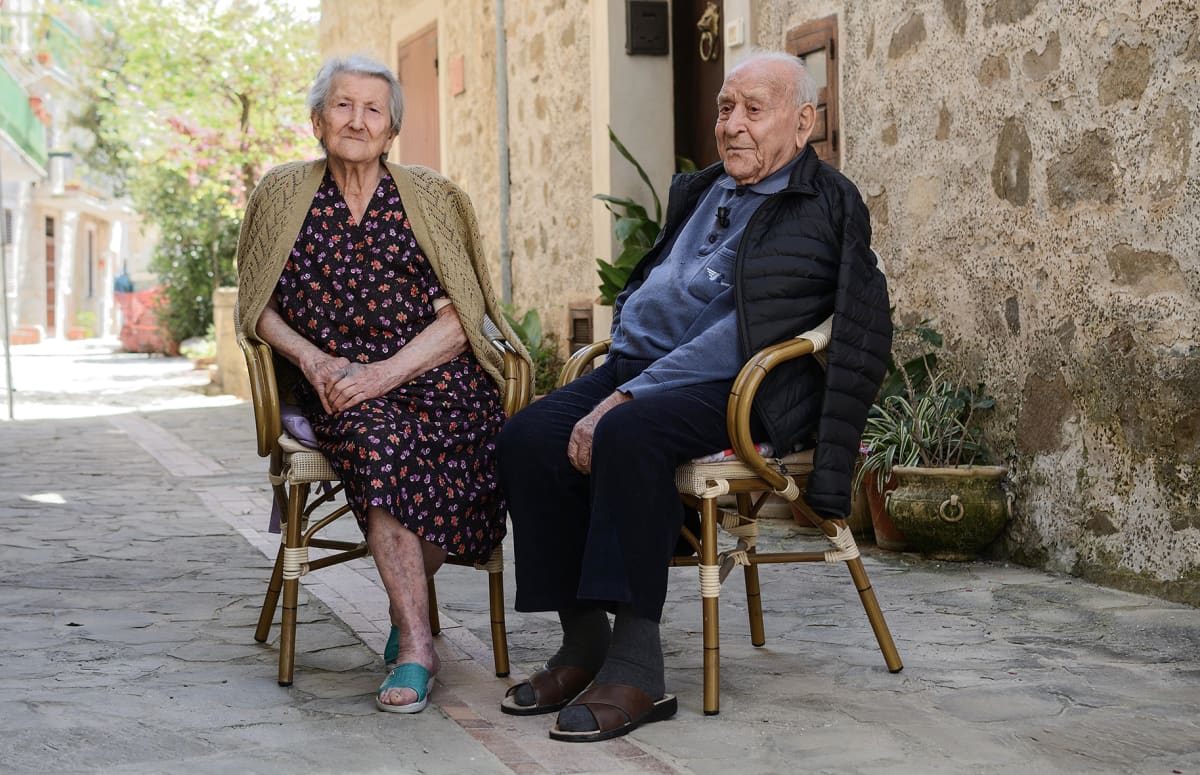 Amina Fedullo ja Antonio Vassallo nauttivat raikkaasta ulkoilmasta asuntonsa edustalla Acciarolin kylässä. Vanha pariskunta istuu tuoleilla.