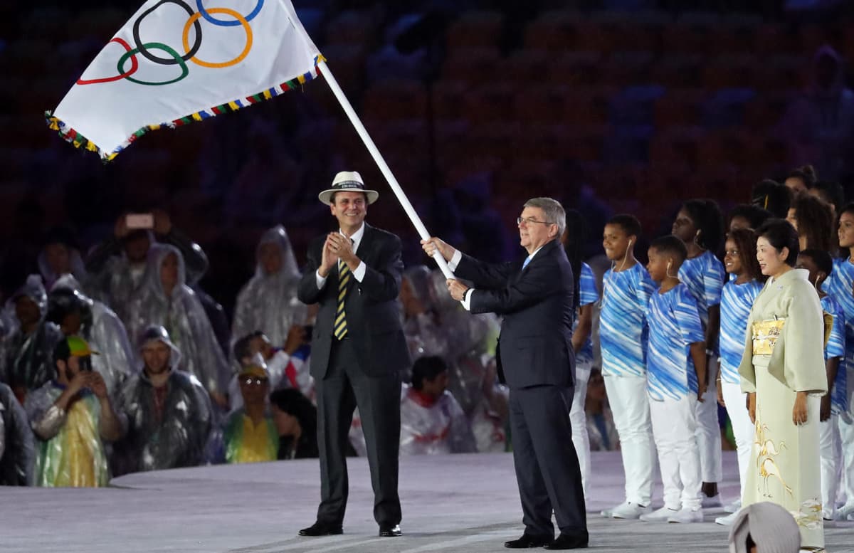 Rion entinen kaupunginjohtaja taputtaa lavalla, kun KOK:n presidentti Thomas Bach liehuttaa olympia-lippua Rion kesäolympialaisten päättäjäisissä. Lavalla on myös tuleva kisaisäntä Tokion kuvernööri Yuriko Koike. 