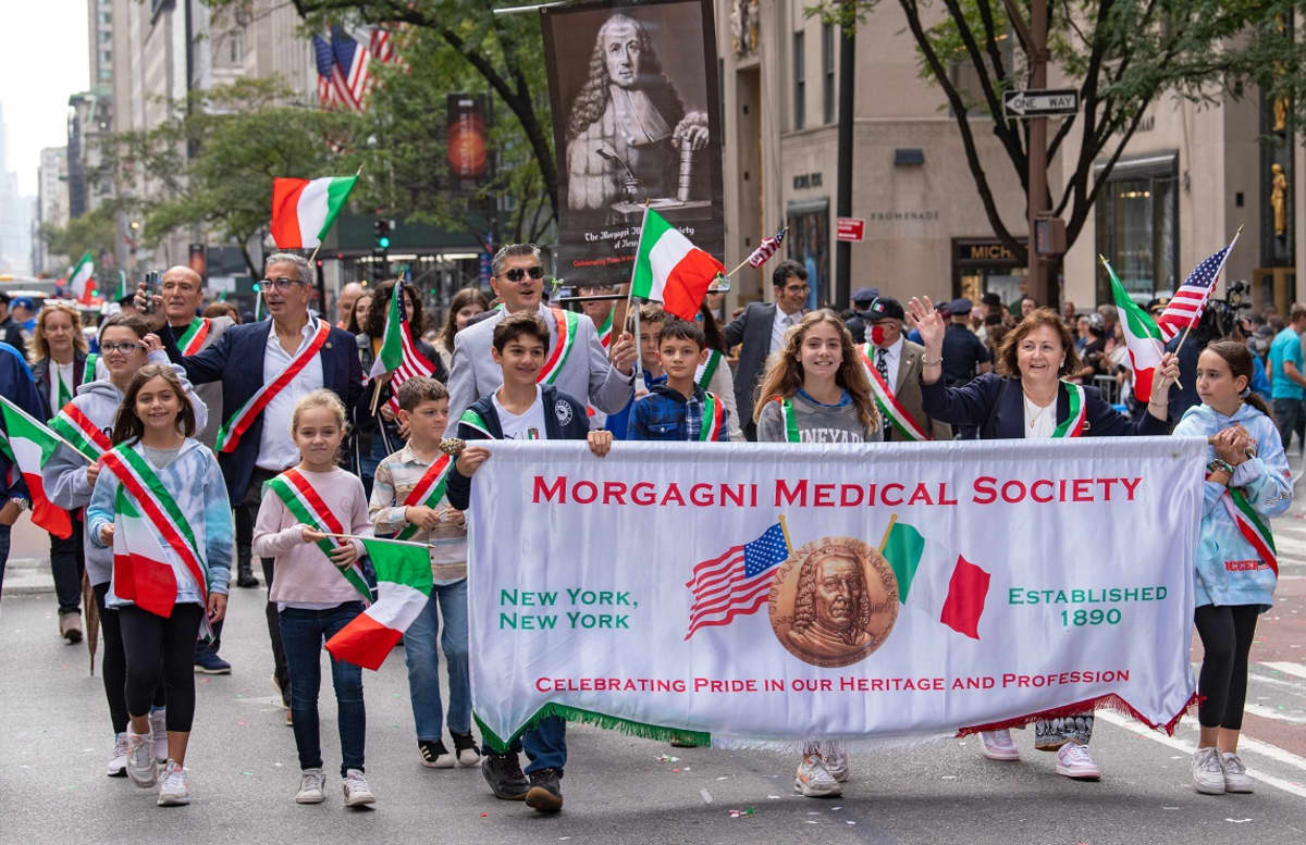 Italian lippuja heiluttelevia ihmisiä, joilla on myös olkanauha Italian lipun väreissä. Eturivissä marssivilla on Morganin lääketieteellisen järjestön banderolli, jossa marssijat kertovat juhlivansa ylpeinä sukujuuriaan ja ammattiaan. 