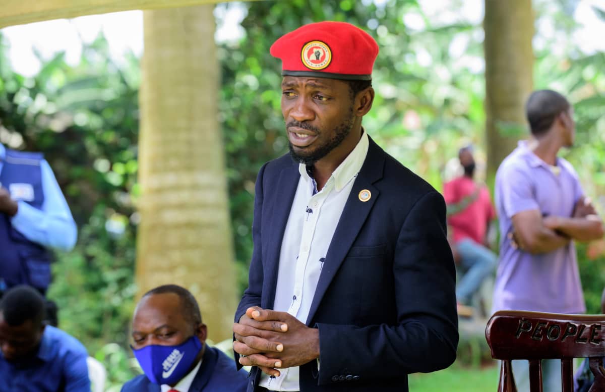 Bobi Wine puhuu tiedotustilaisuudessa ulkotilassa. Hänellä on tumma puku, vaalea kauluspaita ja päässään punainen baretti.