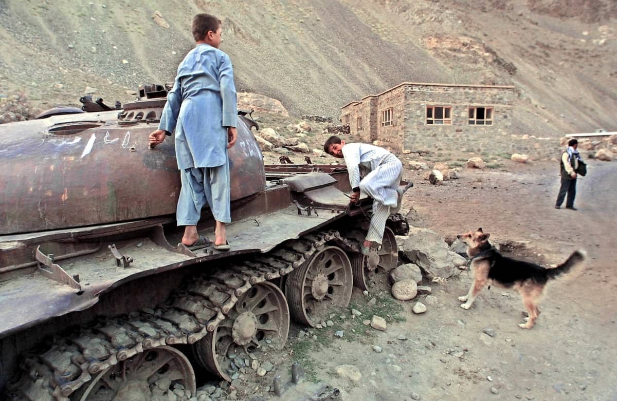 Kaksi poikaa leikkii tuhoutuneen panssarivaunun päällä. Vaunun juurella seisoo koira ja katsoo vaunun päälle nousevaa poikaa. Taustalla näkyy koruttoman näköinen talo ja mies. Talon takana rinne nousee jyrkästi.