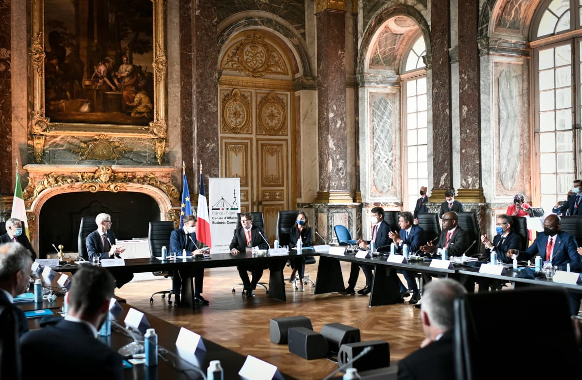 Iso neliönmuotoinen pöytä Versailles'n juhlasalissa, liikemiehiä pöydän ympärillä, Macron keskellä.