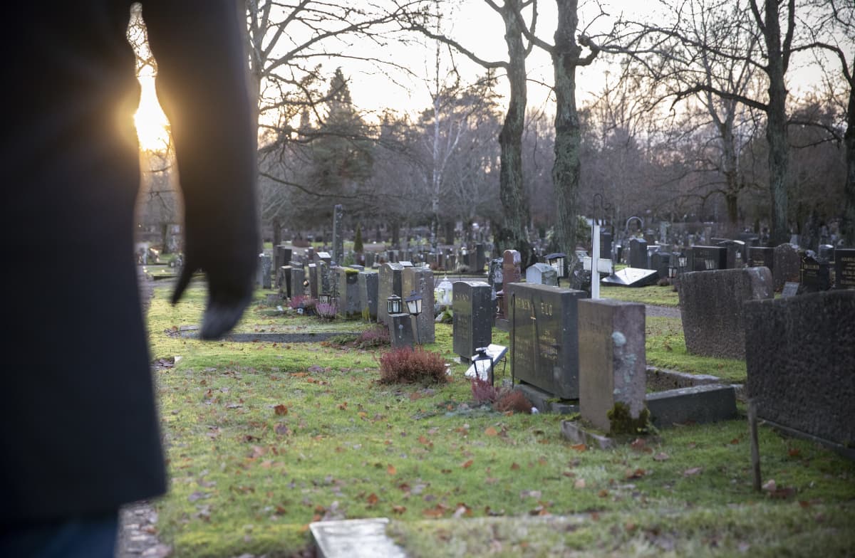 Mies on hautausmaalla ja katselee hautakiviä. Hän seisoo kuvan vasemmassa laidassa ja hänestä näkyy vain osa vartaloa ja oikea käsi. Laskeva talvinen aurinko pilkottaa käden kainalosta.