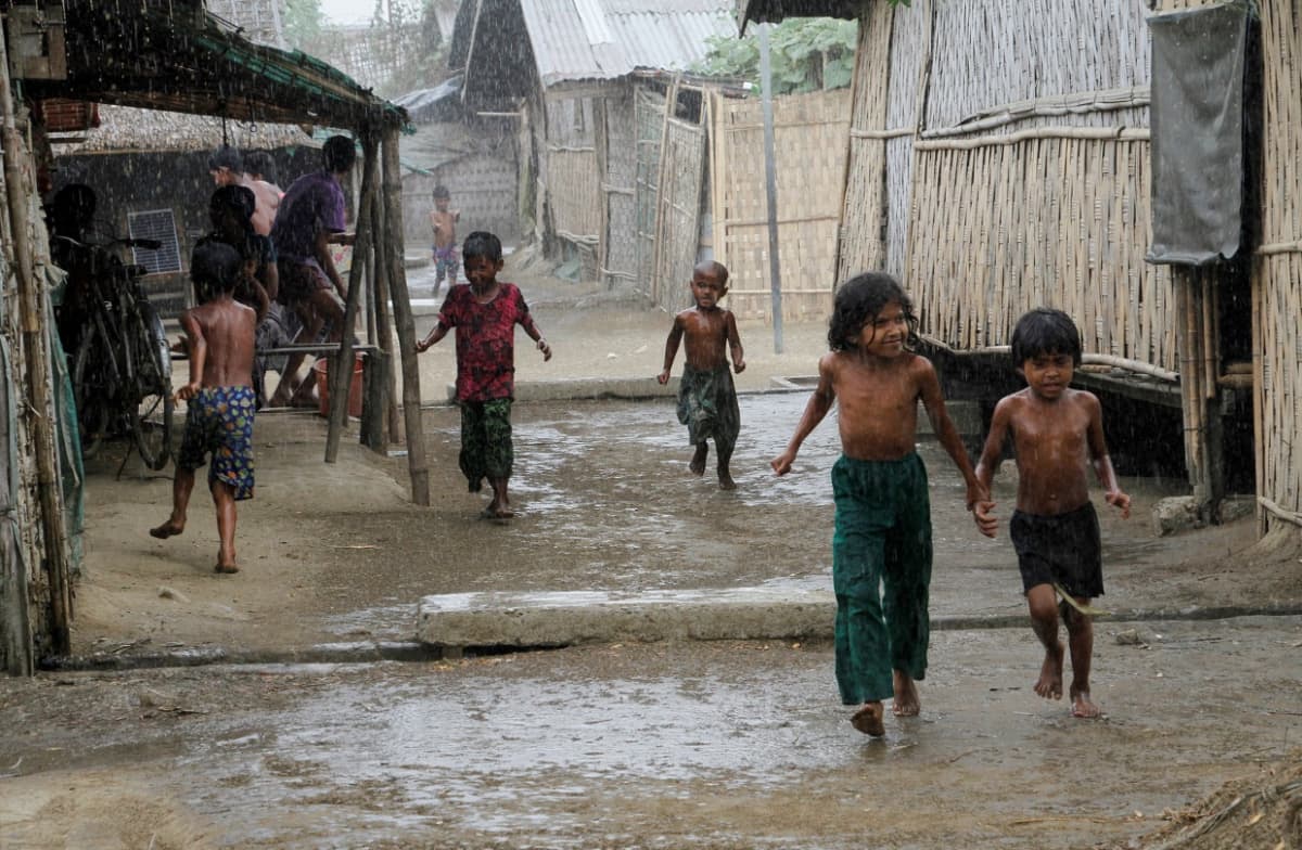 Lapsia juoksee sateessa. Bamburakenteisia taloja kylän raitilla. 