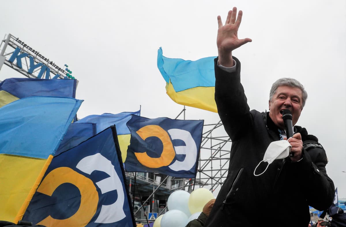 Poroshenko puhuu mikrofoniin ulkona. Hänellä on tumma päällystakki yllään. Poroshenko nostaa oikean kätensä korkealla ilmaan. Taustalla näkyy Ukrainan ja hänen puolueensa Eurooppalaisen solidaarisuuden lippuja sekä Kiovan lentokentän kyltti.