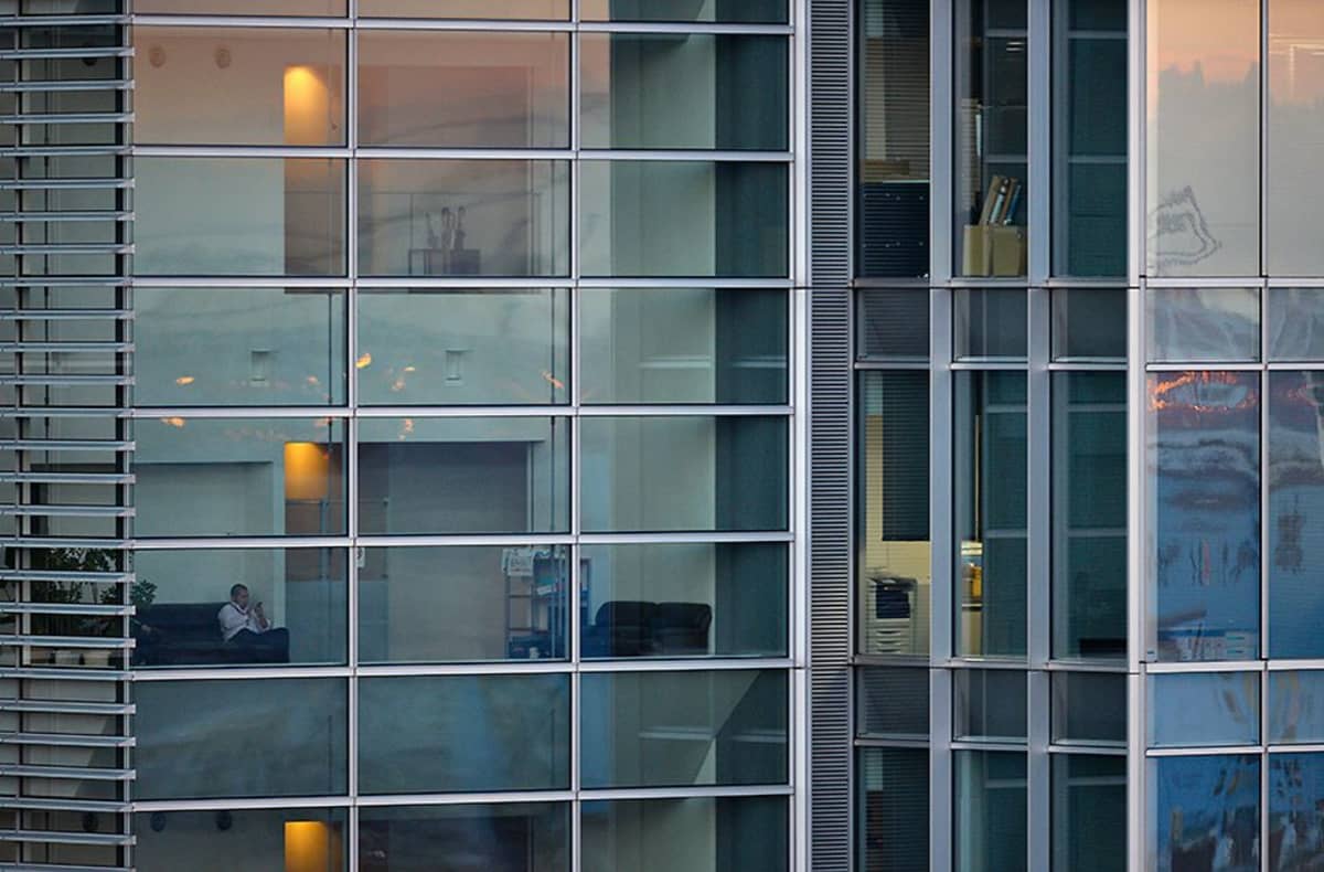 Valkoiseen paitaan pukeutunut japanilaismies istuu sohvalla isossa toimistorakennuksessa Japanin Tokiossa. Aurinko alkaa laskea ulkona ja myös sisätilojen keltaiset valot näkyvät lasisista toimistoikkunoista.