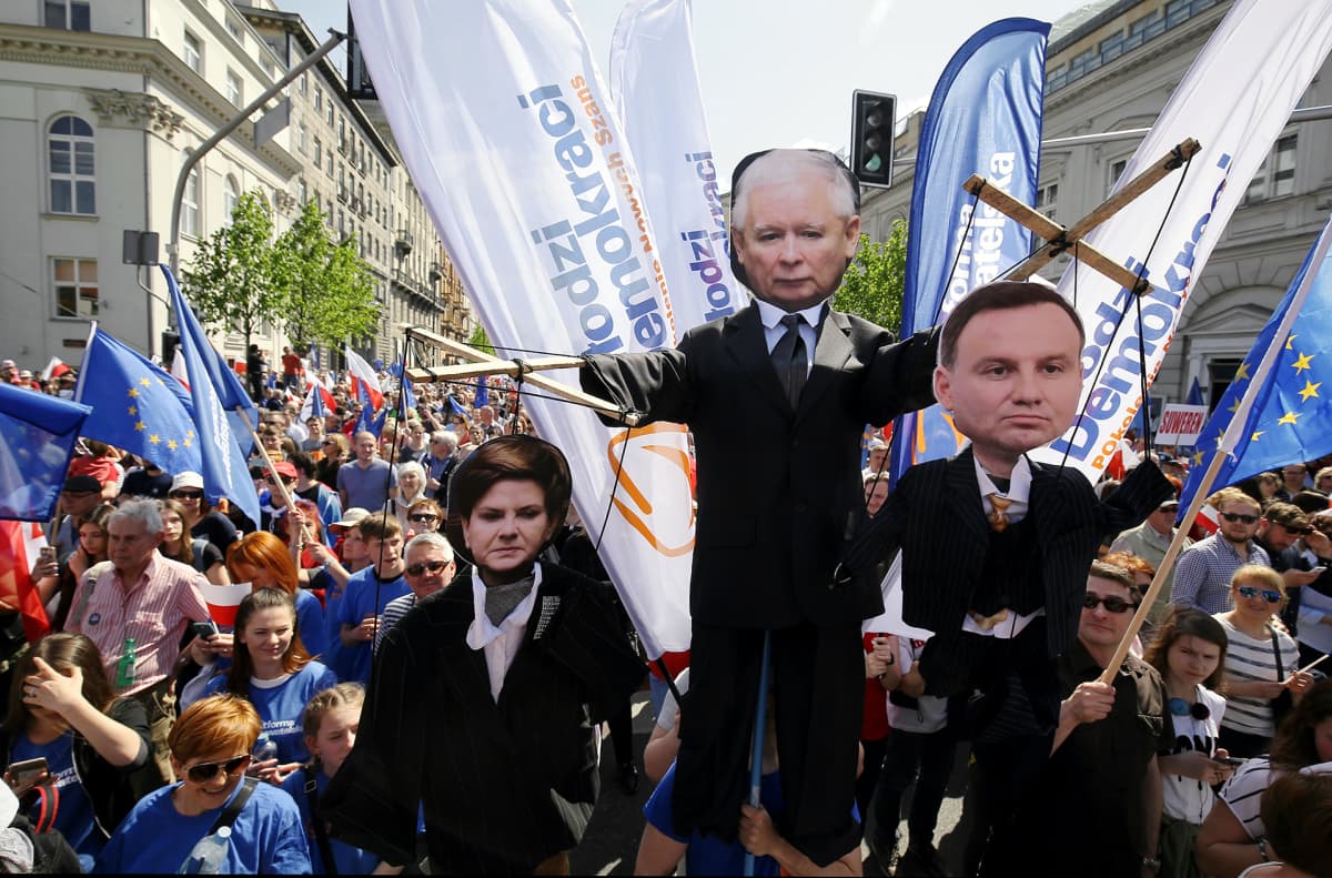 Suuri joukko ihmisiä kulkee katua pitkin. Ihmisillä on käsissään muun muassa EU-lippuja. Etualalla kannetaan isoja nukkehahmoja. Jarosław Kaczyńskia esittävä nukke riiputtaa pääministeriä ja presidenttiä esittäviä nukkeja naruista.