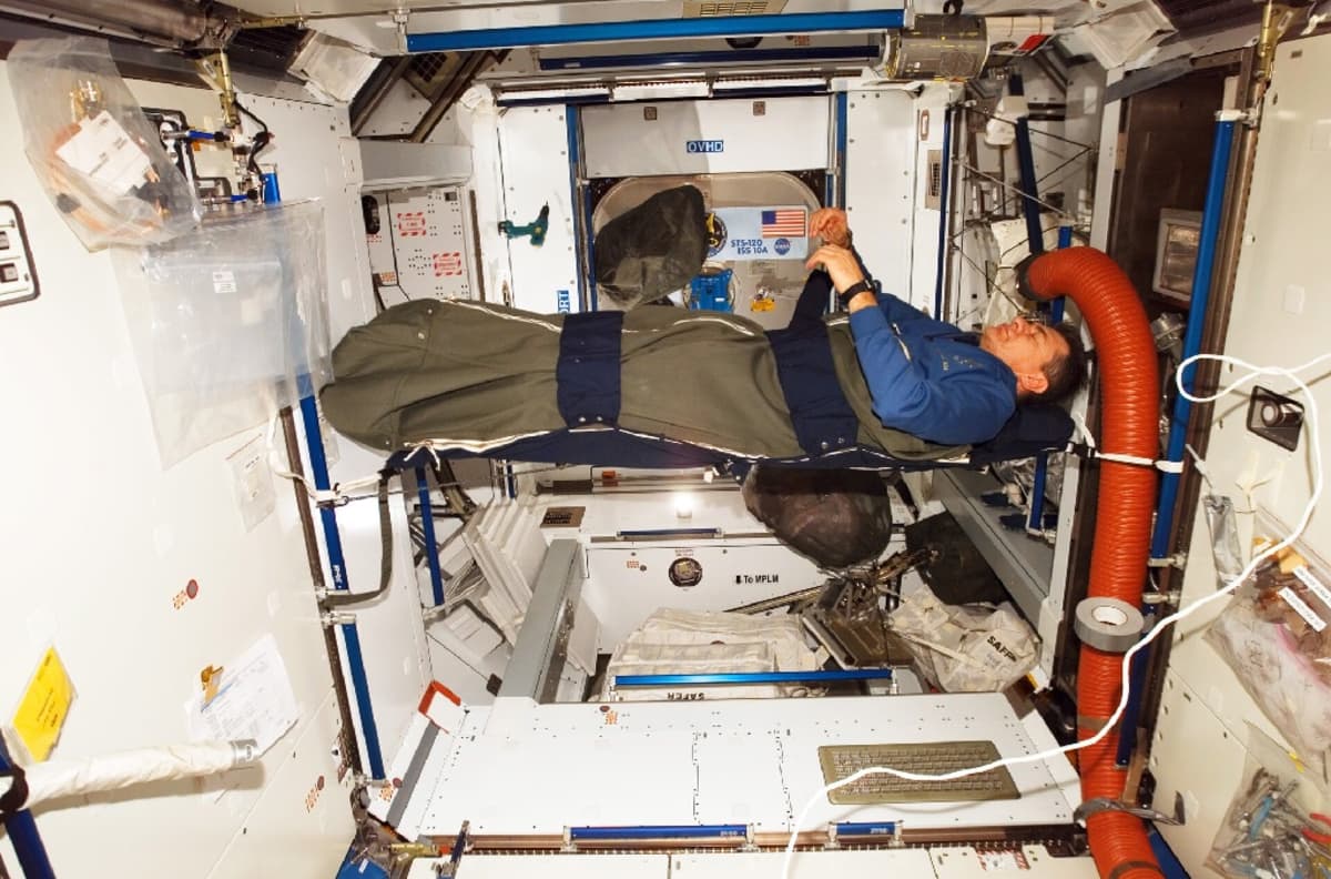 Mies makuulla avaruusaseman rakenteisiin vöillä kiinnitetyssä makuupussissa. Hänen kätensä leijuvat ilmassa.  