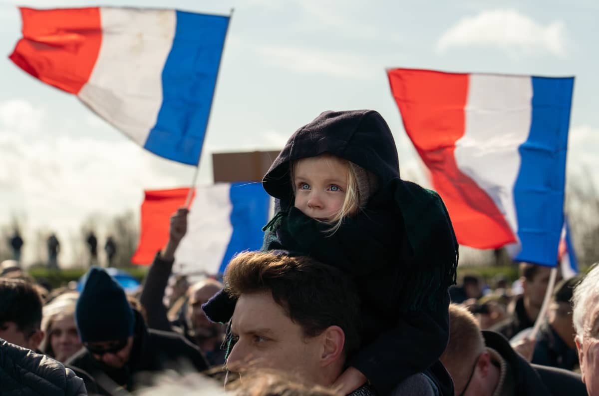 Lapsi istuu miehen hartioilla yleisön keskellä. Taustalla näkyy liehuvia Ranskan lippuja.