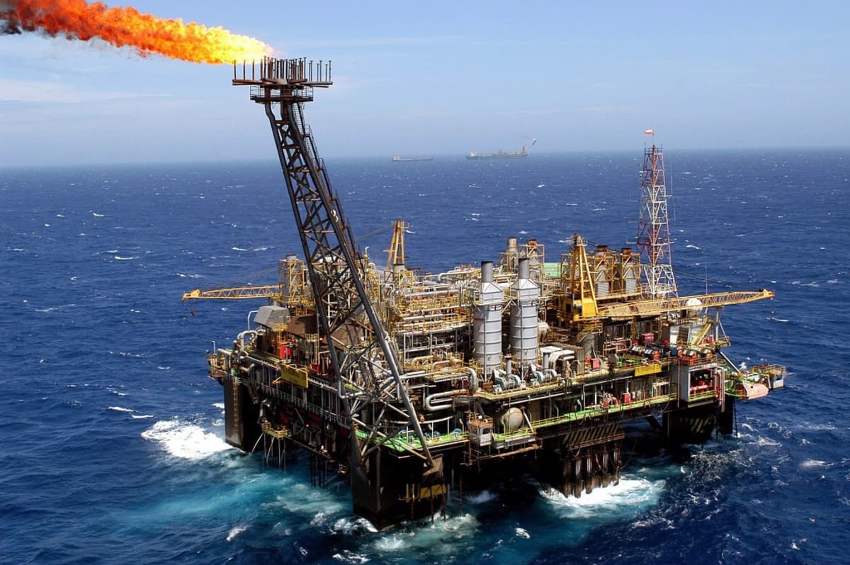 Brasilian Petrobras-yhtiön öljylautta merellä päivällä. Meri aaltoilee tummansinisenä, taivas on vaalea. Öljynporauslautan tornista nousee liekki, kun porauksen yhteydessä nousevaa kaasua poltetaan. Kaukana horisontissa näkyy kaksi alusta, mahdollisesti tankkereita.