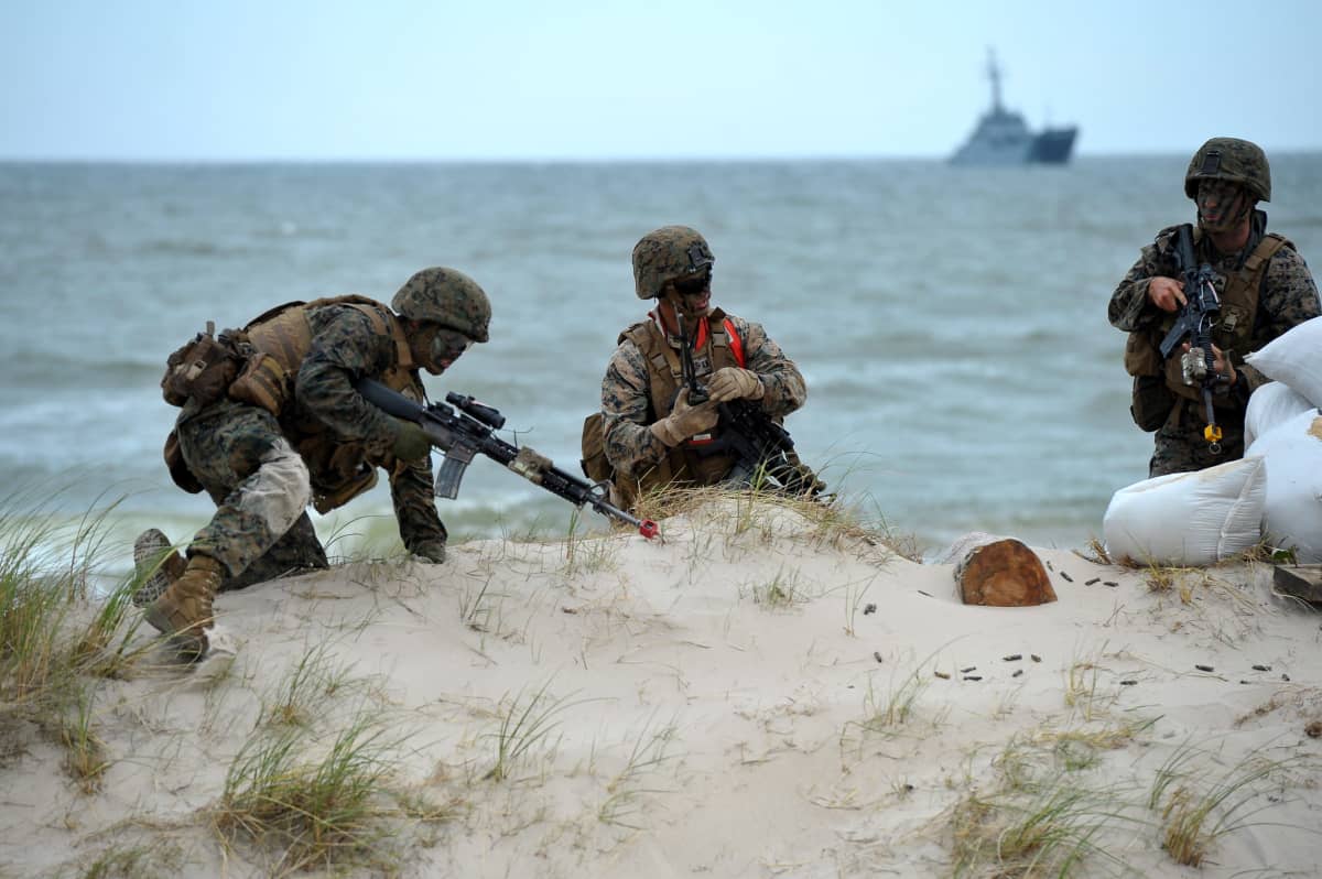 Sotilaita hiekkatöyräällä meren rannalla ja laiva takana merellä.