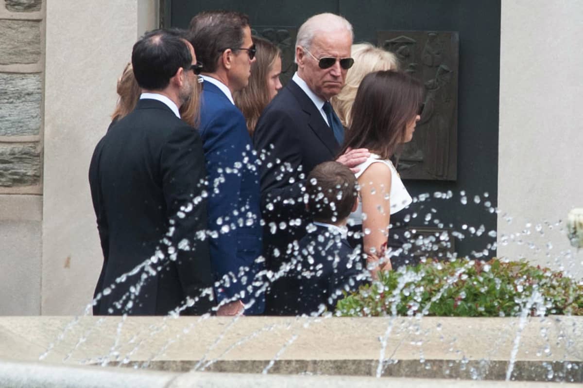 Joe Biden perheineen Bidenin esikoispoika Beaun hautajaisissa. Kuvan etualalla on suihkulähde. Biden perheineen on kuvassa tiiviisti yhdessä. Bidenillä itsellään on mustat aurinkolasit ja hän on ainoa joka katsoo kuvaajaa kohden, ilme vakavana.