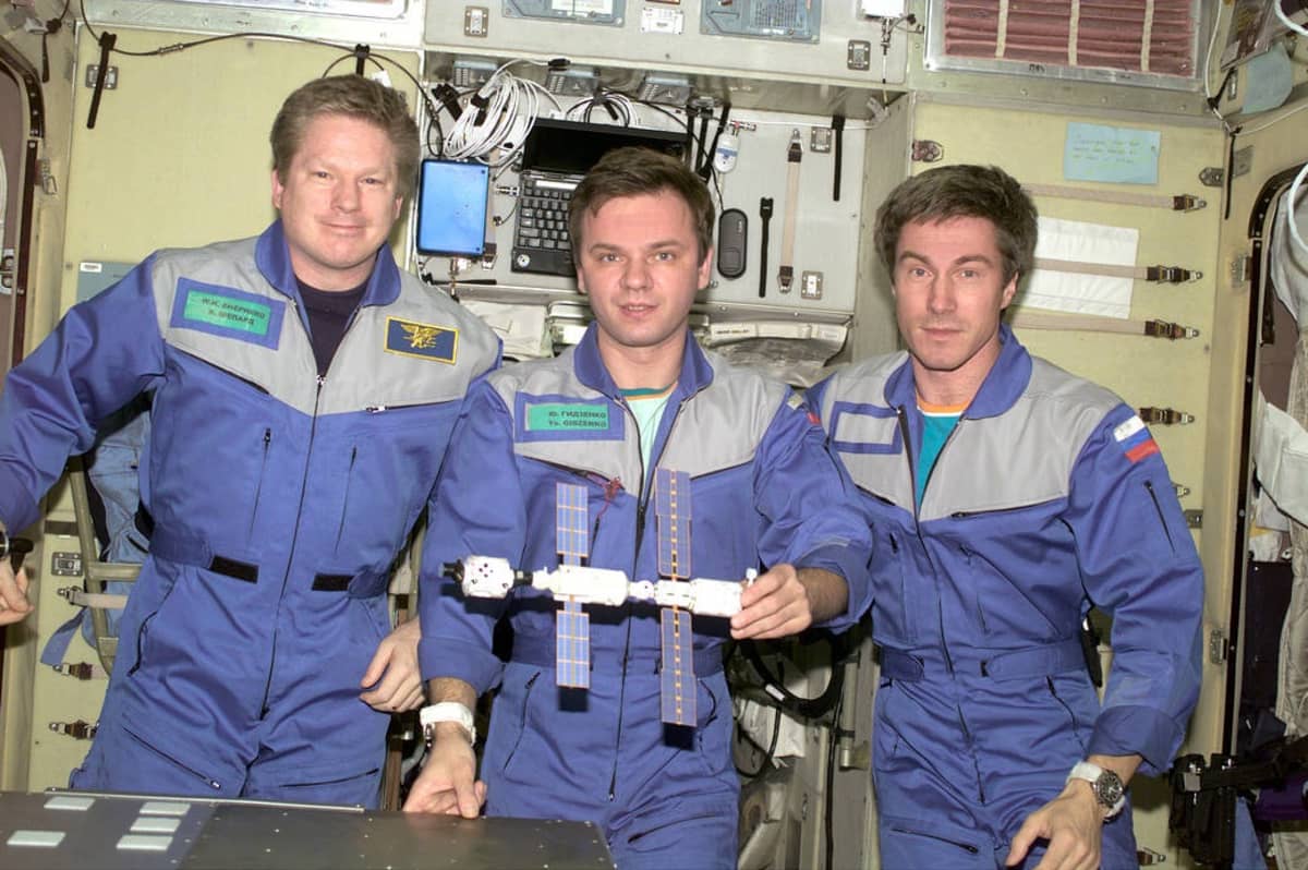Kolme miestä sinisissä kokohaalareissa, taustalla avaruusaseman laitteistoa. 