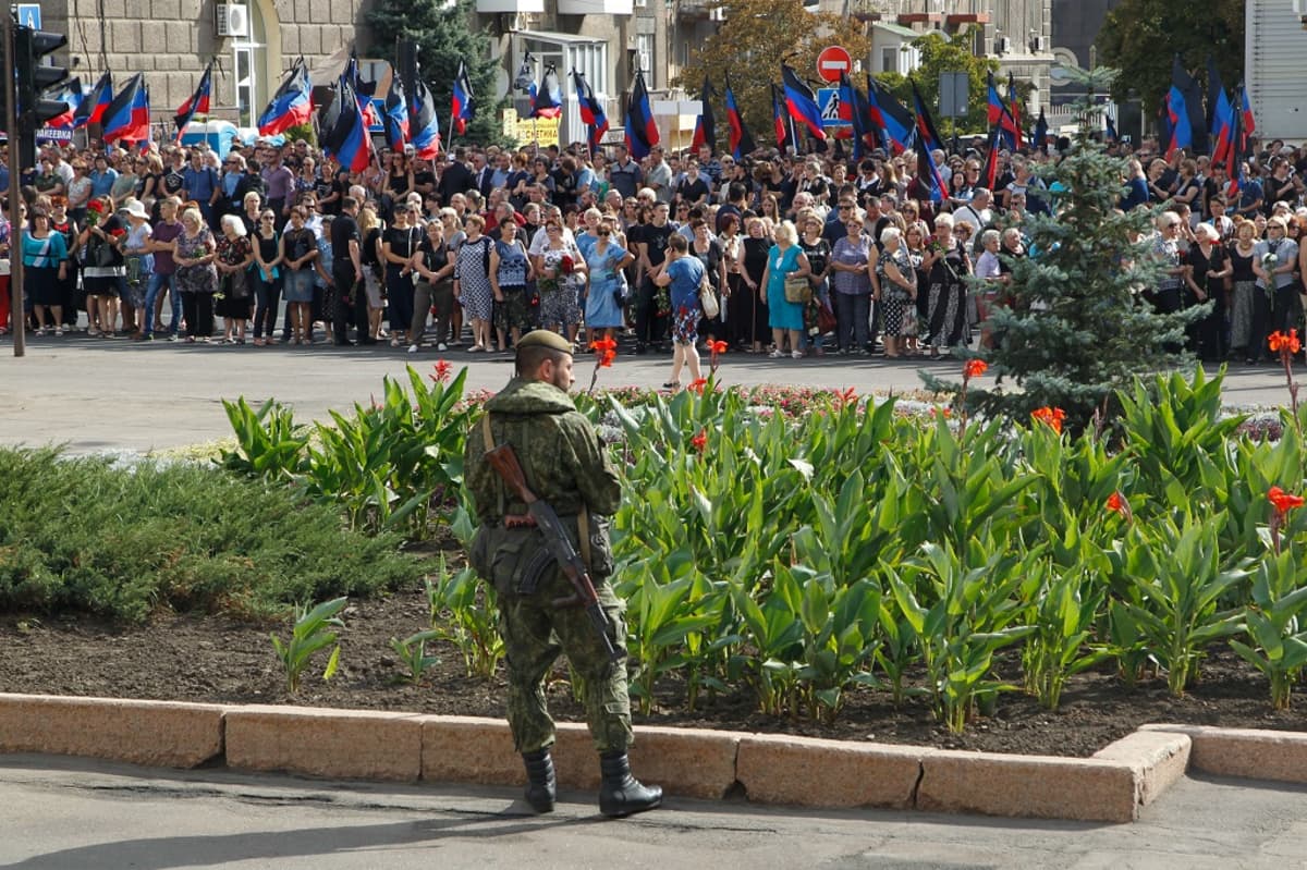 Väkijoukko seisoo kadun varrella odottamassa Aleksandr Zahartšenkon hautajaissaattoa. Väkijoukossa näkyy Donetskin kapinallisalueen musta-sini-punaisia lippuja. Etualalla kadun toisella puolella kukkaistutusten edessä seisoo maastounivormuun pukeutunut parrakas mies baretti päässään ja rynnäkkökivääri selässään.
