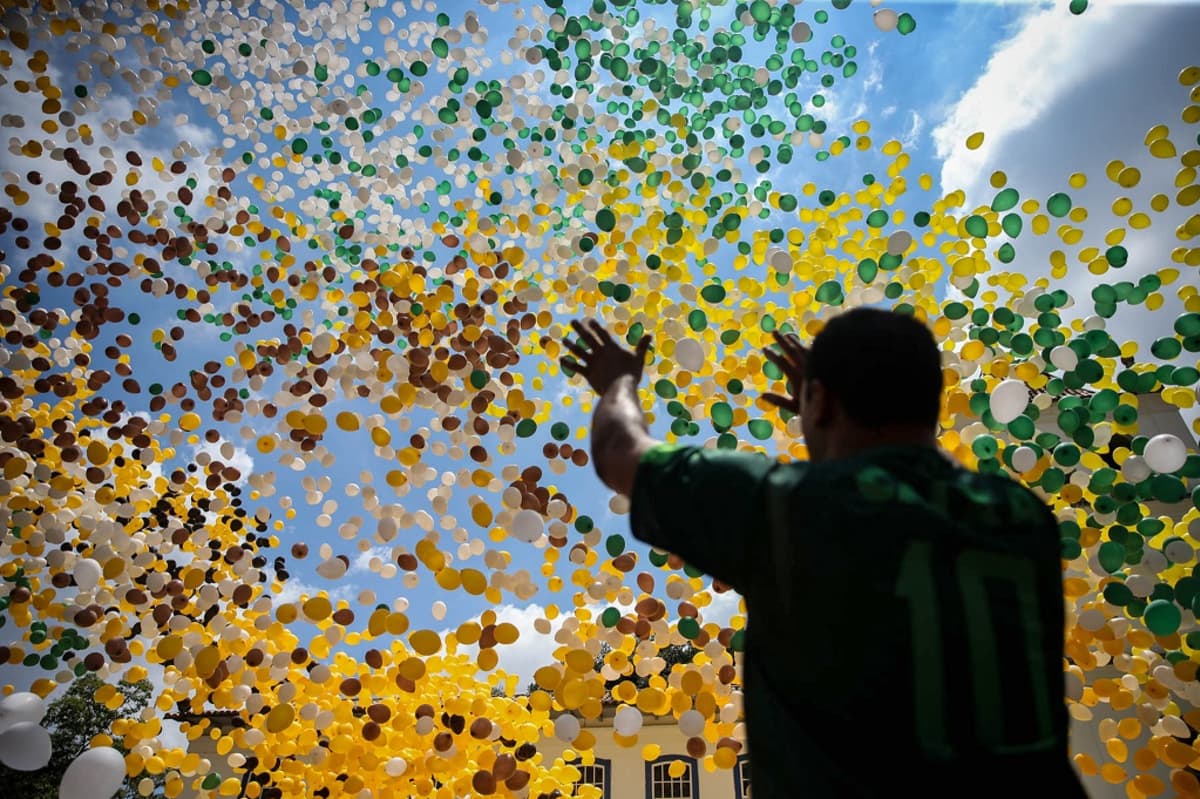 Mies ojentaa käsiään kohti taivaalle kohoavaa Brasilian lipun väristen ilmapallojen suurta parvea. 