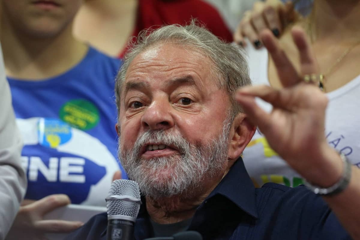 Parrakas Lula da Silva puhuu oikeassa kädessään olevaan mikrofoniin ja elehtii vasemmalla kädellään, jonka etusormi ja peukalo ovat yhdessä. Lulalla on tummansininen kauluspaita. Taustalla seisoo kannattajia.