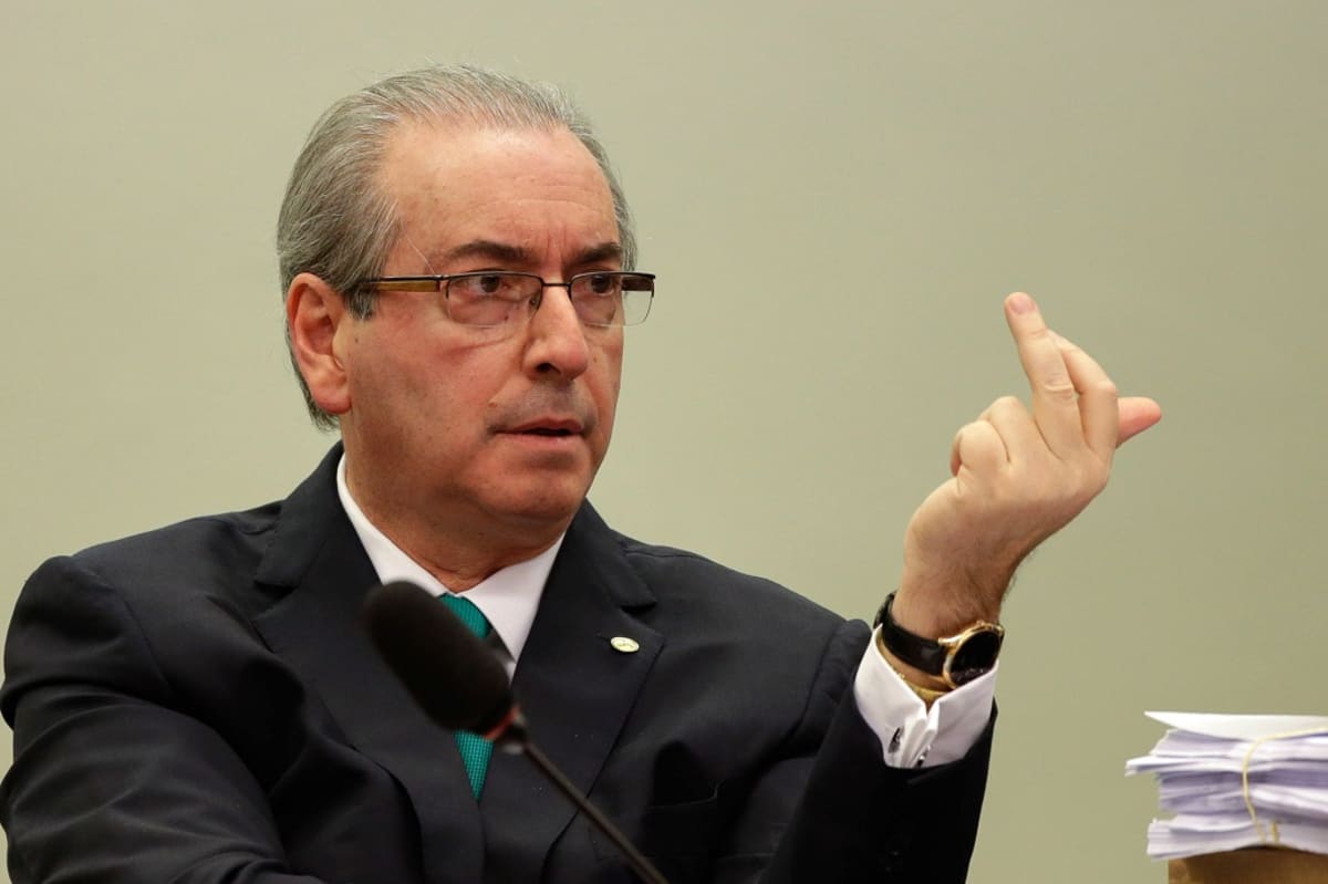 Eduardo Cunha istuu mikrofonin takana ja pitää vasenta kättään pystyssä.