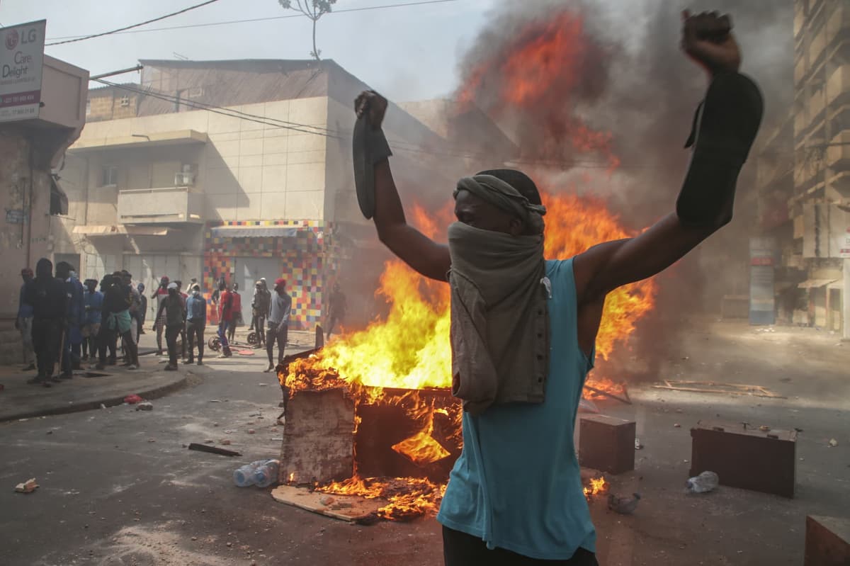 Mielenosoittaja seisoo kädet ylhäällä palavien esineitten edessä Dakarissa.