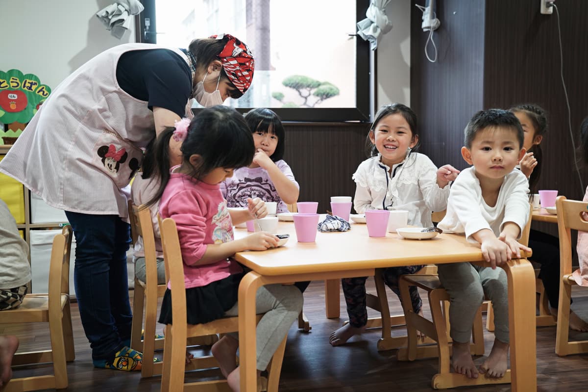 Lapsia syömässä pöydän ääressä, kasvosuojusta käyttävä hoitaja kumartuu pöydän ylle.