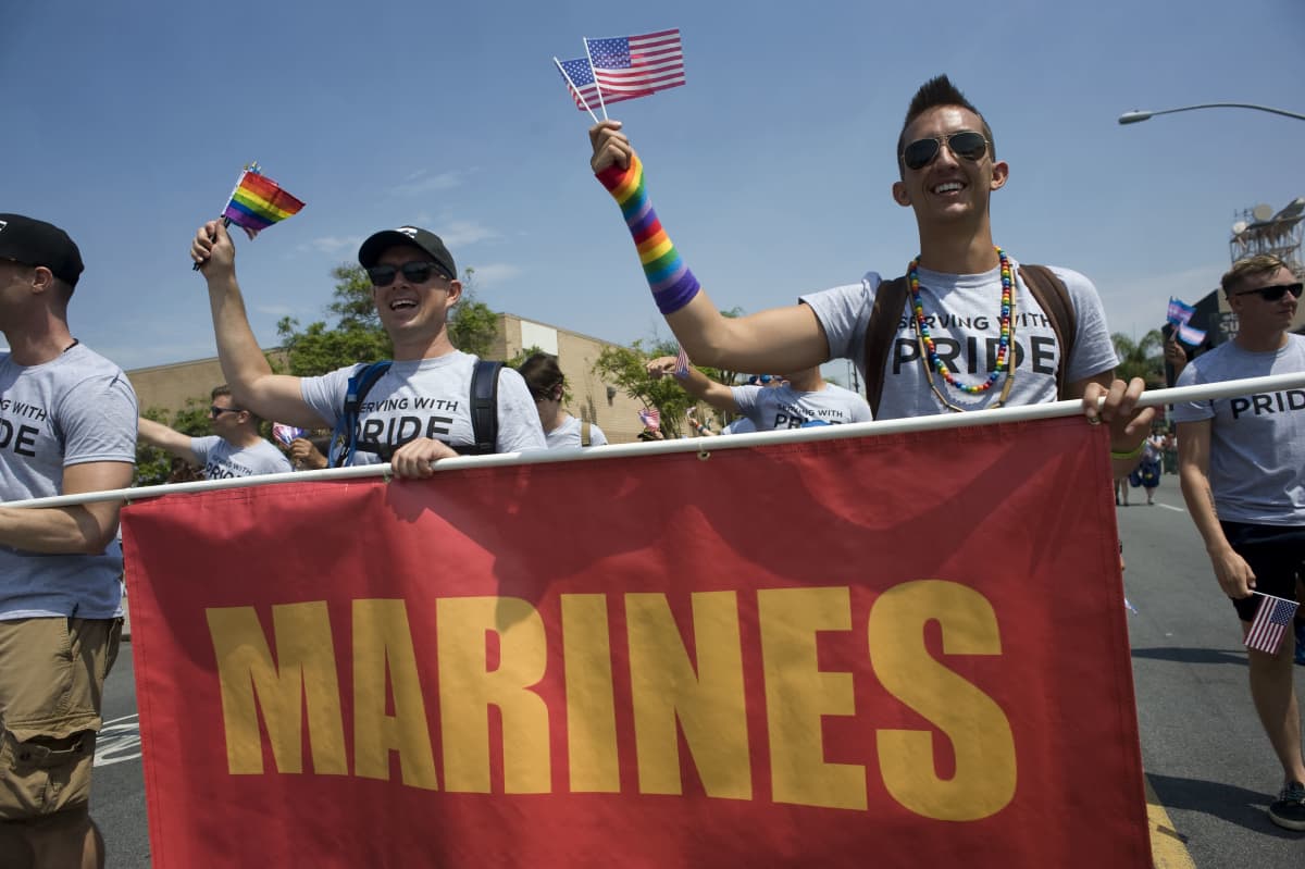 Kaksi miestä marssii pride-kulkueessa ja heiluttaa Yhdysvaltain- ja sateenkaarilippua. He kantavat kylttiä, jossa lukee "marines".
