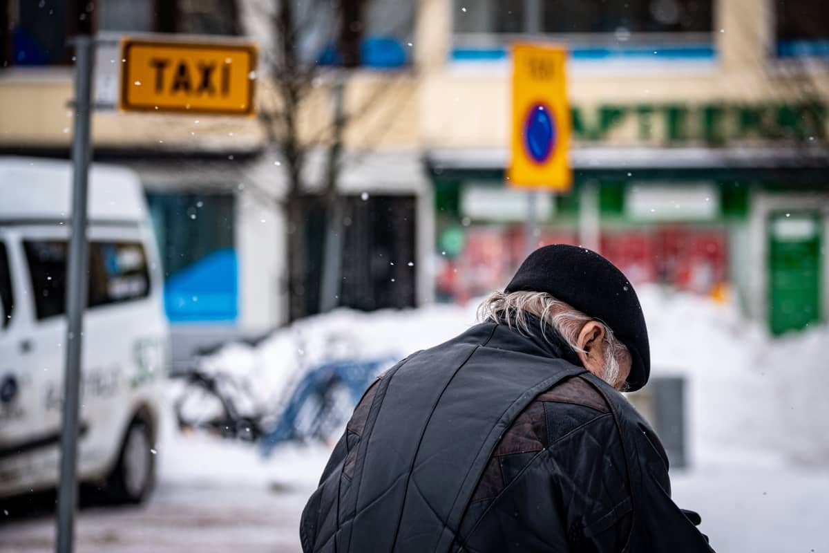 Pysäköity taksi ja vanhempi mies talvisessa kaupunkimaisemassa.