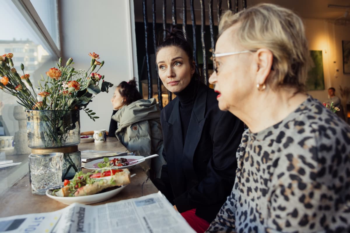 Noora Hautakangas, yrittäjä, Relove second hand -vaatekaupan ja kahvilan omistaja ja vakioasiakas Liisa Zilliacus (oik.) istuu pöydän ääressä ja katsoo ikkunasta ulos. 