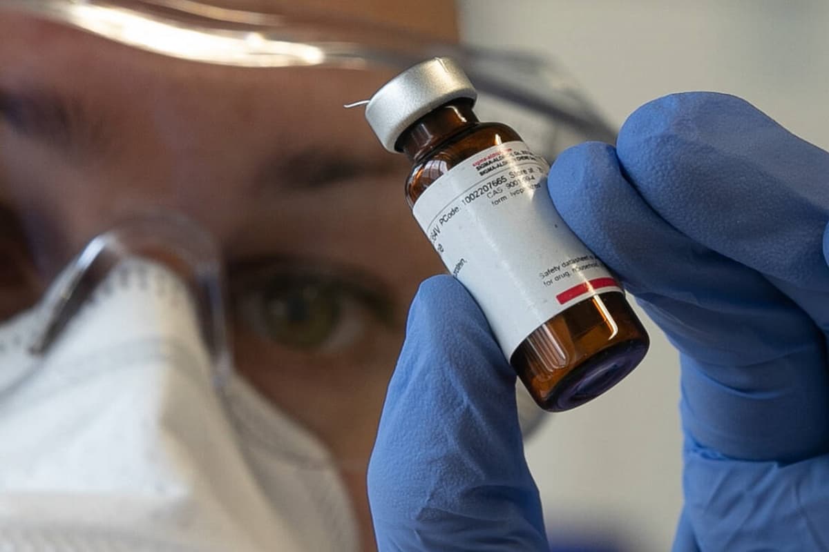 Lääkepullo ja pipetti suojavarusteisiin pukeutuneen tutkijan käsissä. 