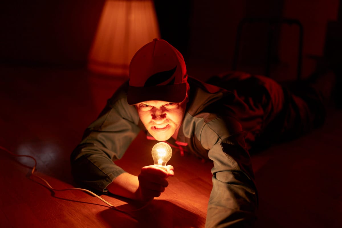 Näyttelijä Juho Uusitalo makaa lattialla ja pitää hehkulamppua kädessään kuin mikrofonia. Hänellä on työhaalari ja lippis.