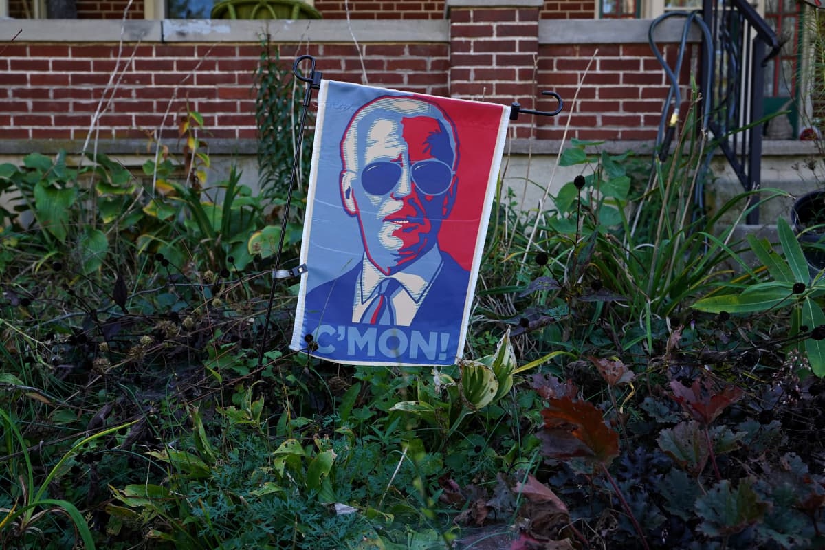 Joe Bidenia kannatta kyltti puutarhassa. Punavalkosinisessä kyltissä on Bidenin kuva ja teksti C'mon!.