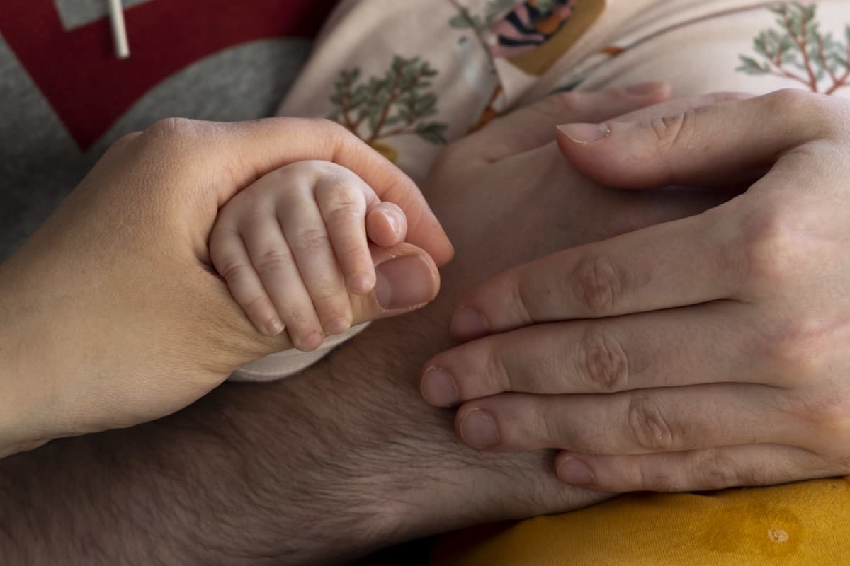 Aikuinen pitää vauvan kättä kädessään. Kaksi aikuisen karvaista kättä pitelee vauvaa sylissä. 