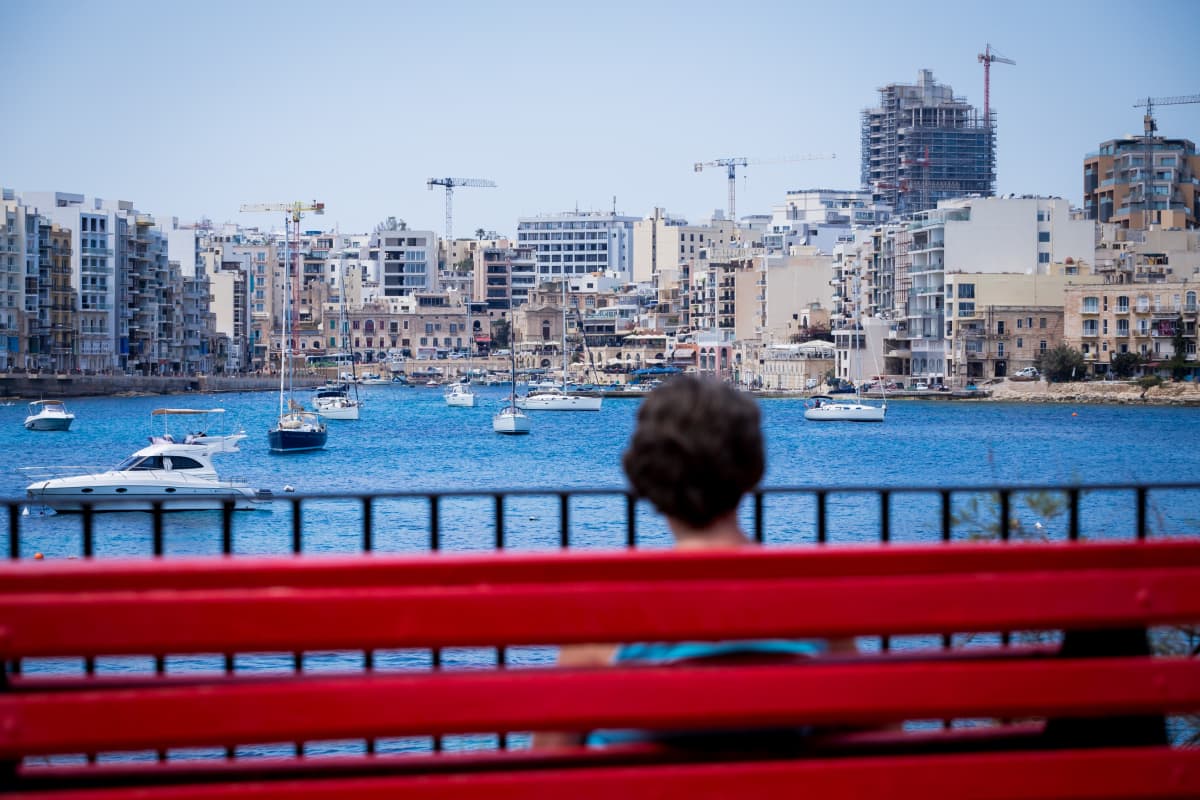 Nainen istuu penkillä rannassa, takana kaupunkisiluetti, jossa nostokurkia