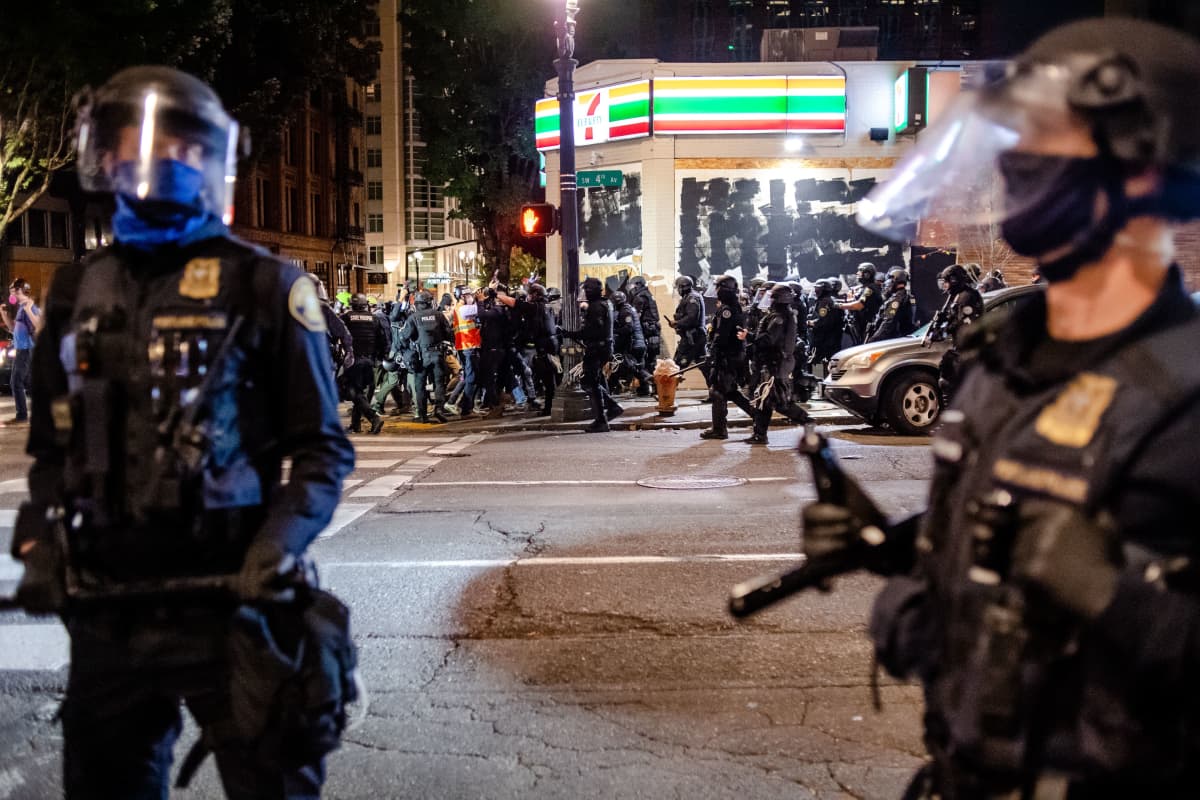 Iltavalaistuksessa kaupunkikuvassa näkyy mellakavarusteisia poliiseja ja taustalla mielenosoittajia. 