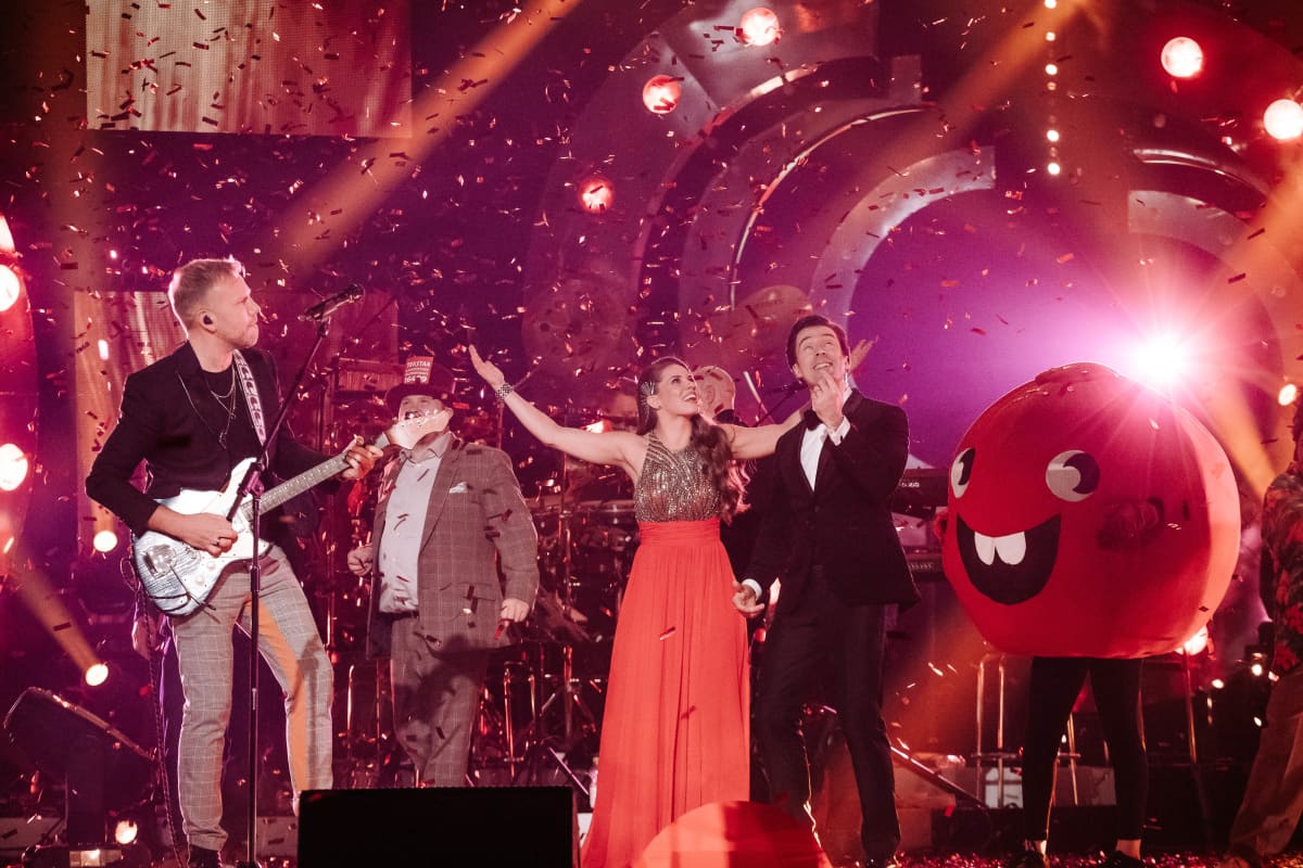 Nenäpäivän juontajat iloitsevat rahankeräyspotista muusikoita taustallaan.