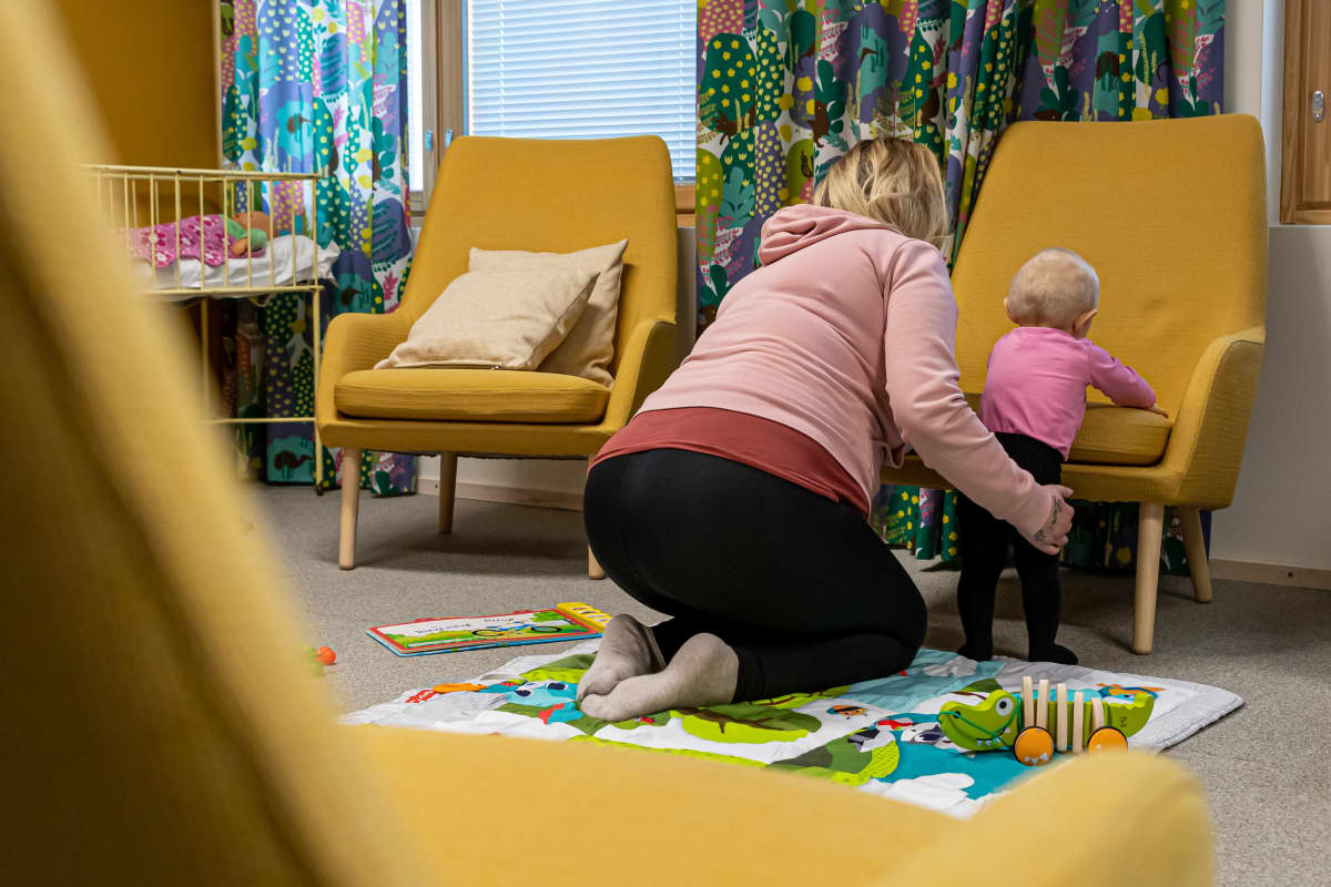 Alle vuoden ikäinen lapsi seisoo lattialla nojatuolin istuinta vasten. Hänen vieressään istuu lapsen äiti polvillaan ja pitää hellästi kiinni.