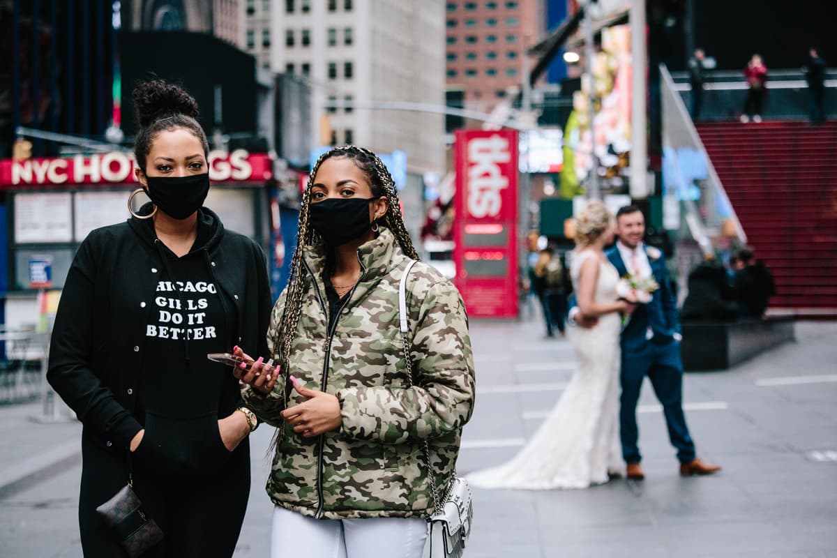 Kaksi naista etualalla pukeutuneena hengitysmaskeihin ja taustalla näkyy hääpari joista otetaan hääkuvia.
