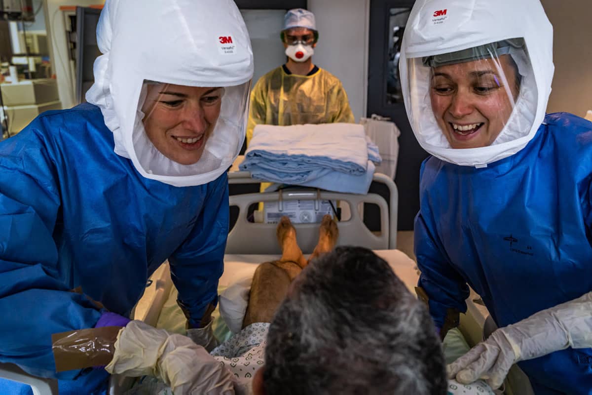 Valokuvateos, jossa kaksi potilasta hoitavaa naista hymyilevät potilaalle. 
