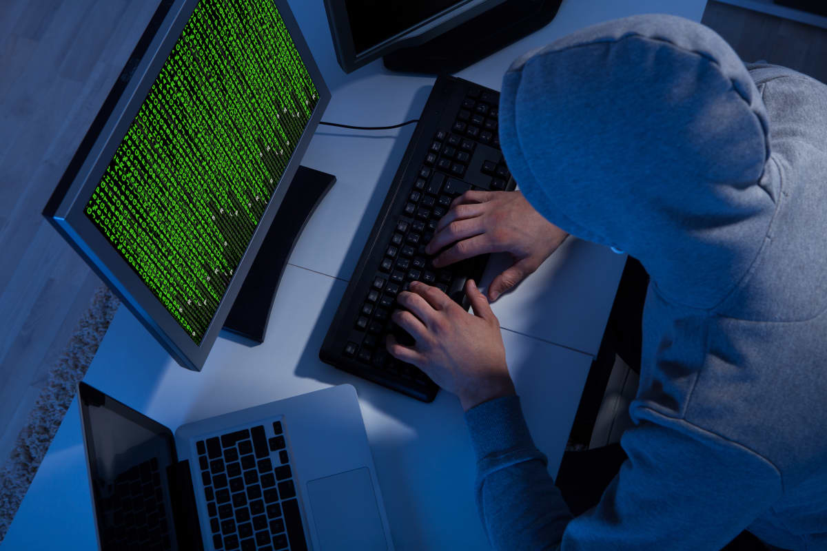 Person iklädd munkjacka sitter vid en laptop och en dator med en massa gröna tecken på skärmen.