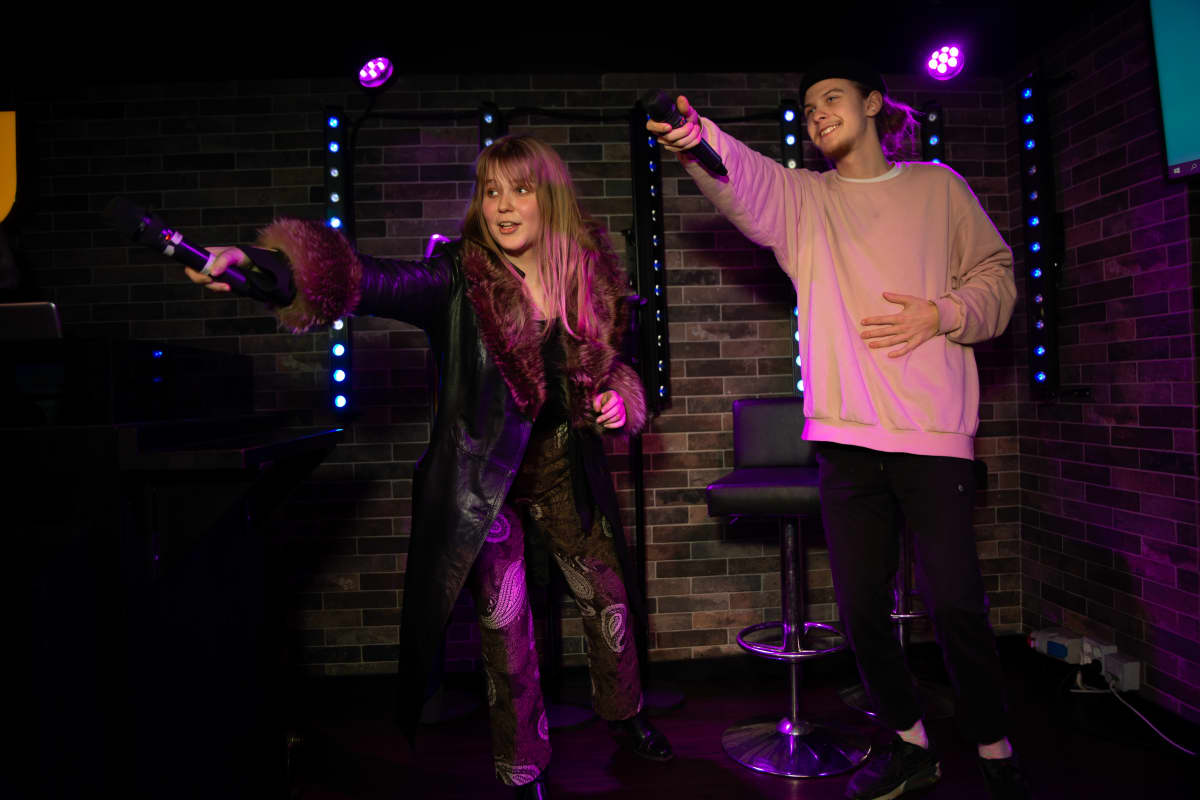 Anna Niemelä ja Olavi Tanttu laulavat karaokea. Kappaleessa on kertosäe meneillään ja laulajat osoittavat mikrofoneillaan kohti yleisöä kannustaen osallistumaan lauluun.