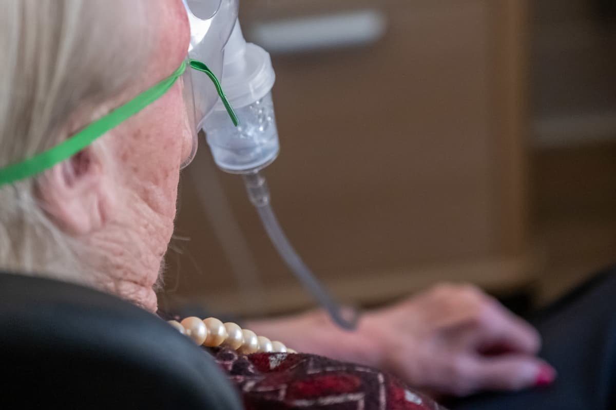 Lähikuva vanhuksesta takaapäin. Vanhuksella happimaski, jolla hän ottaa keuhkolääkettä.
