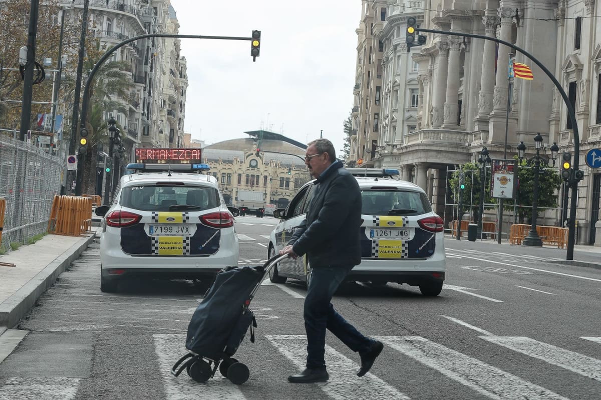 Mies kulkee suojatiellä vedettävän ostoslaukun kanssa. Muita ihmisiä ei näy, vain poliisiautoja.