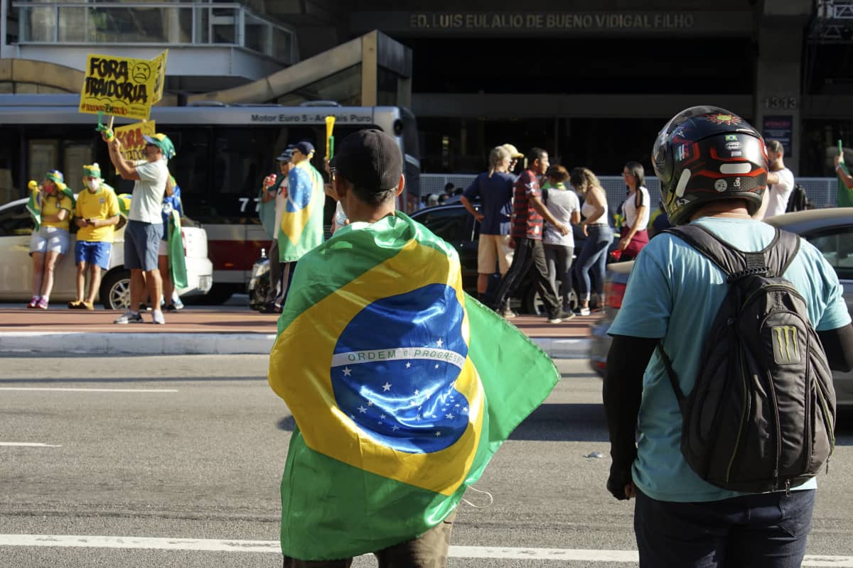 Mielenosoittajia kadulla, lähinnä kameraa oleva henkilö kietoutunut Brasilian lippuun