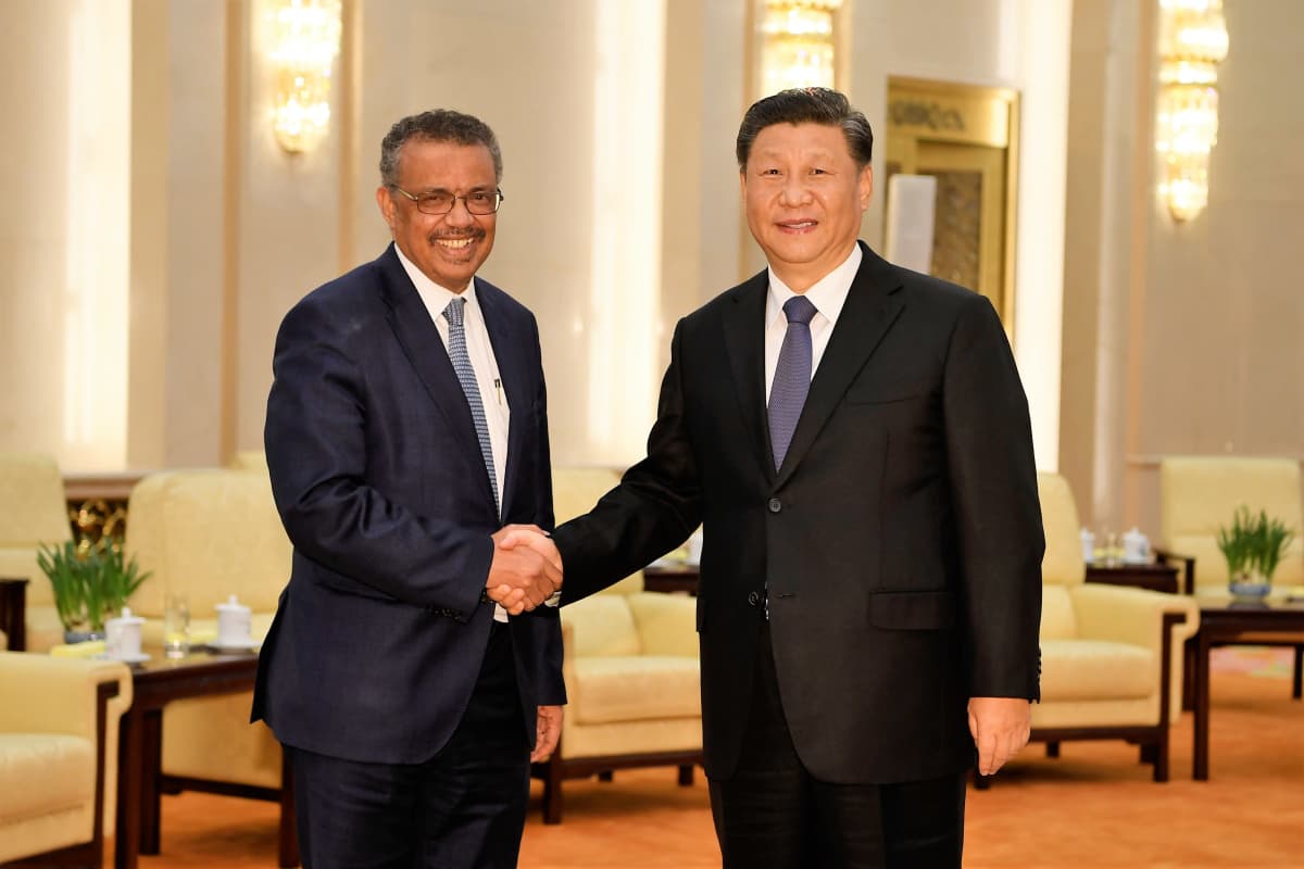 WHO:n pääjohtaja ja Kiinan presidentti kättelevät hymyillen. Miehillä on tummat puvut ja siniset kravatit. Taustalla näkyy salia.