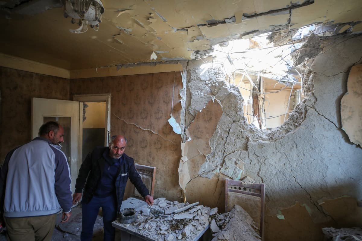 Kaksi miestä tutkii tuhoja huoneessa, jonka seinässä on valtava pommin tekemä aukko.