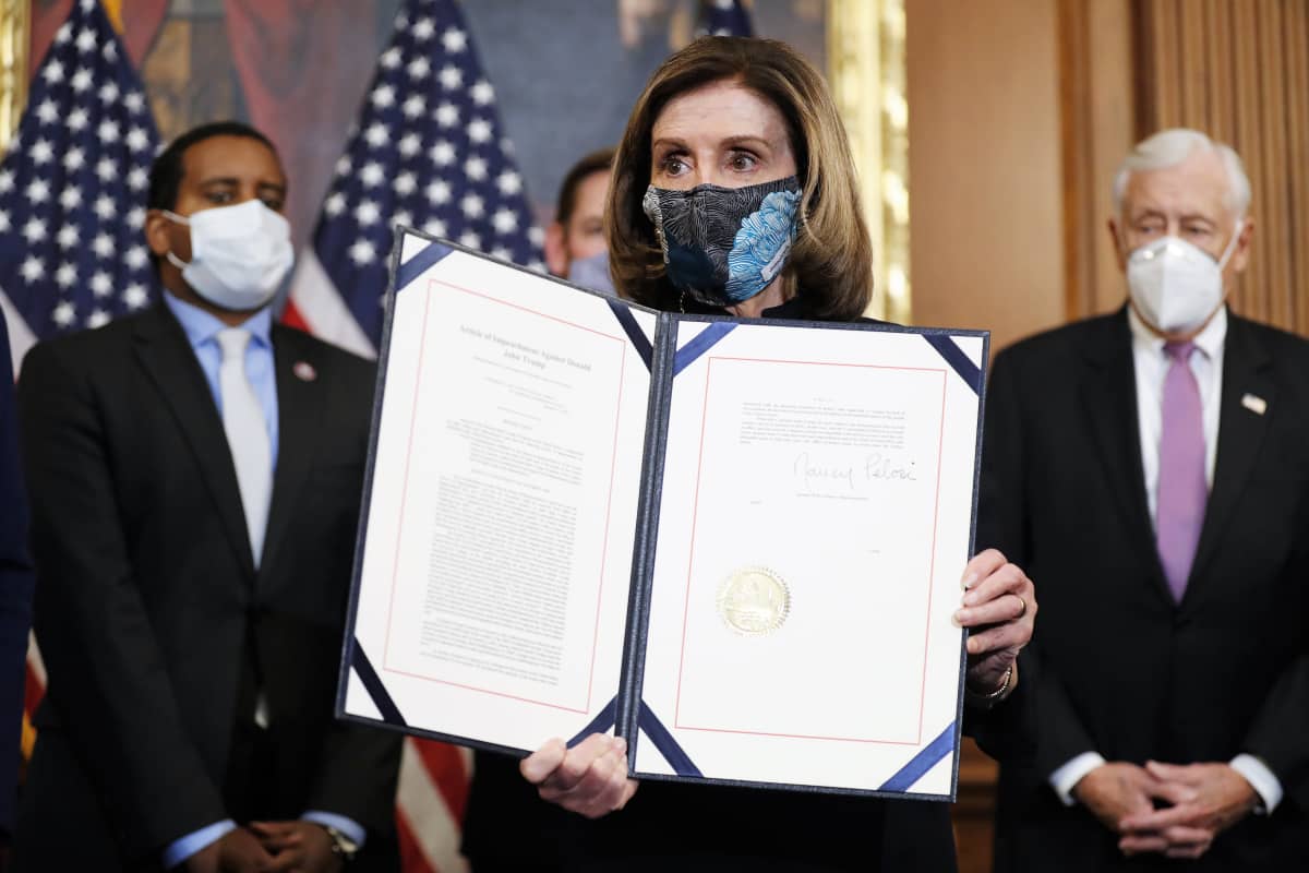 Nancy Pelosi pitää käsissään asiakirjaa ja esittelee sitä kameroille. Hänellä on kasvoillaan maski. Taustalla näkyy kolme miestä tummissa puvuissa ja maskit kasvoilla. Huoneen seinän vierellä on Yhdysvaltain lippuja.