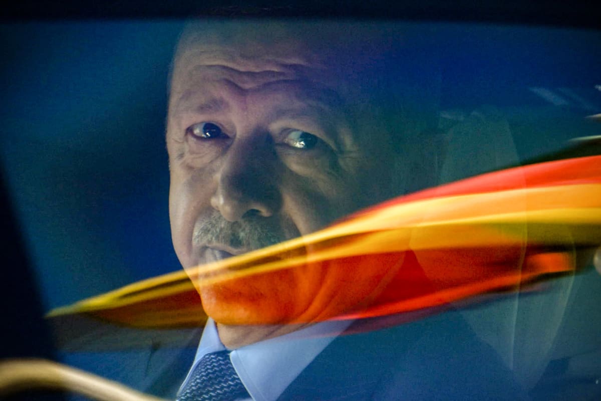 Pääministeri Recep Tayyip Erdogan katsoo suoraan kameraan, välissä on lasi jossa näkyy värillisiä heijastuksia.