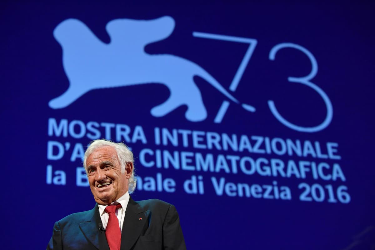 Belmondo seisoo elokuvajuhlien lavalla tummassa puvussa ja punaisessa kravatissa.