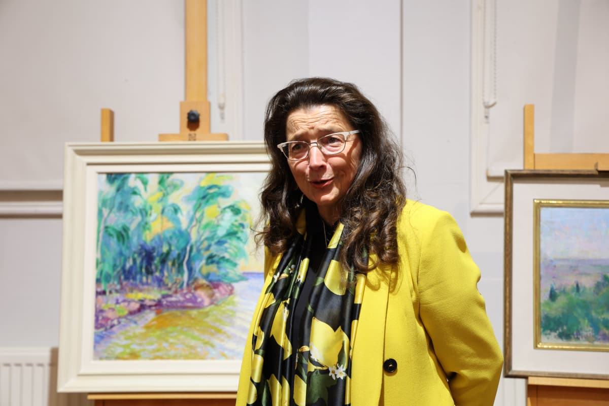 Keltaiseen jakkuun pukeutunut LähiTapiola Idän hallituksen varapuheenjohtaja Mariaana Pykäläinen kertoo Lähellä -näyttelyn teoksista. Taustalla näkyy värikäs maalaus, johon kuvattu koivumetsää ja vesistöä.