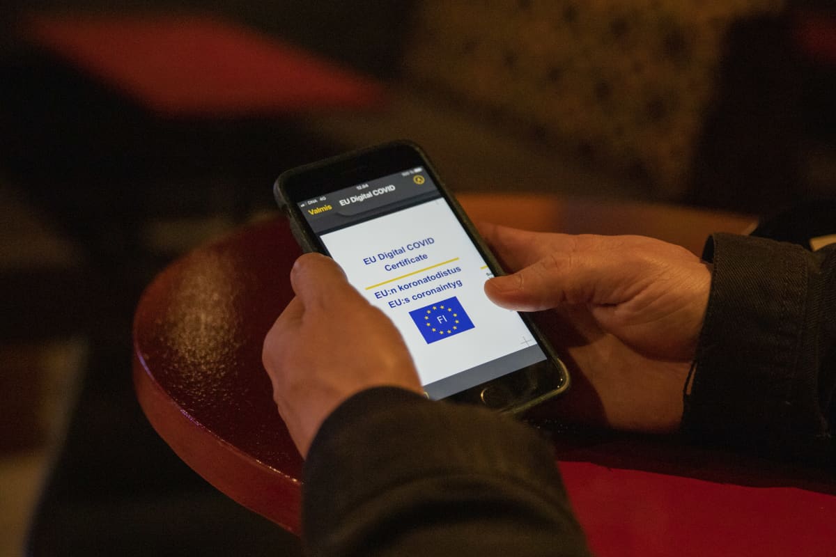 pöydällä olevat kädet pitelevät älypuhelinta, jonka näytöllä näkyy osa koronatodistuksesta.