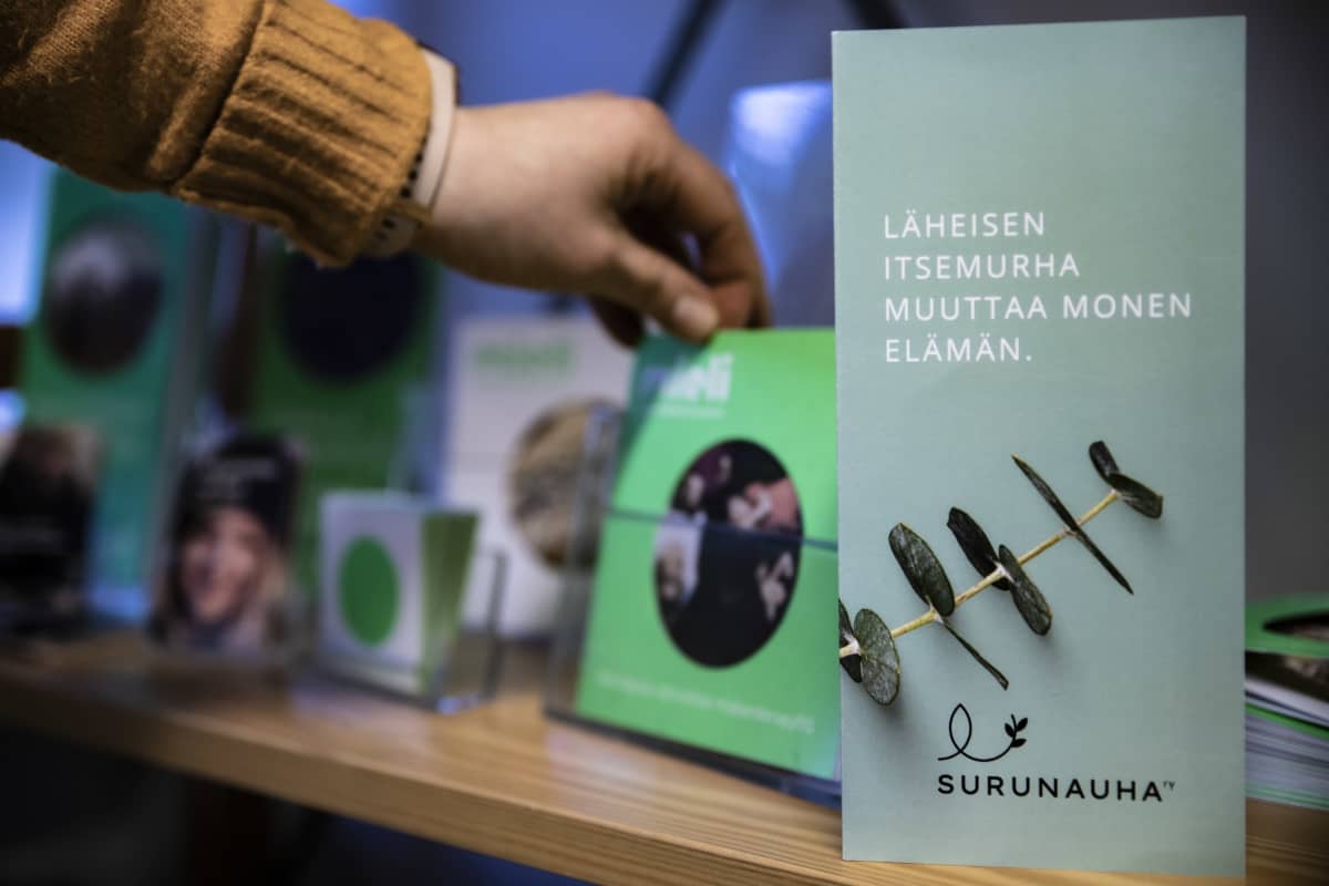 Etualalla Surunauha ry:n esite avusta, jota on tarjolla läheisensä itsemurhassa menettäneelle. Taustalla käsi ottaa toista esitettä.