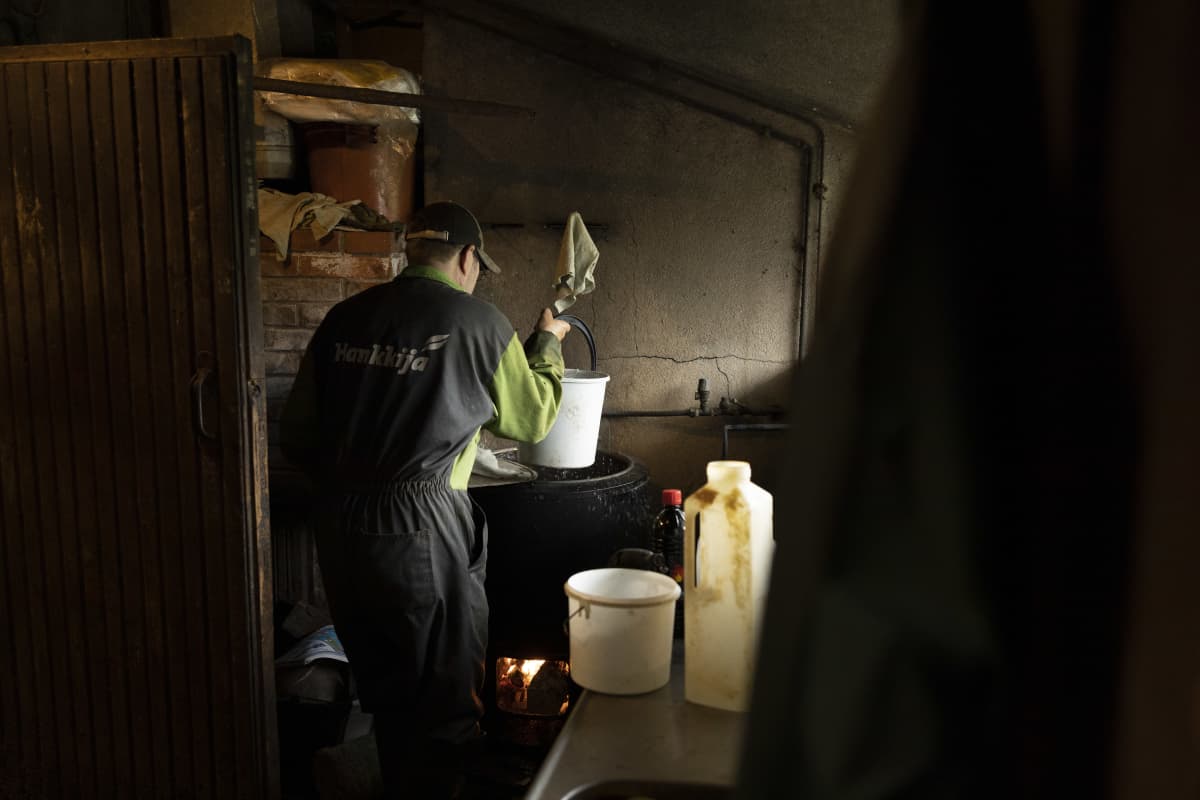 Timo Liimatainen nostaa karjakeittiössä ämpärillä kuumaa vettä padasta, jonka alla palaa tuli. 